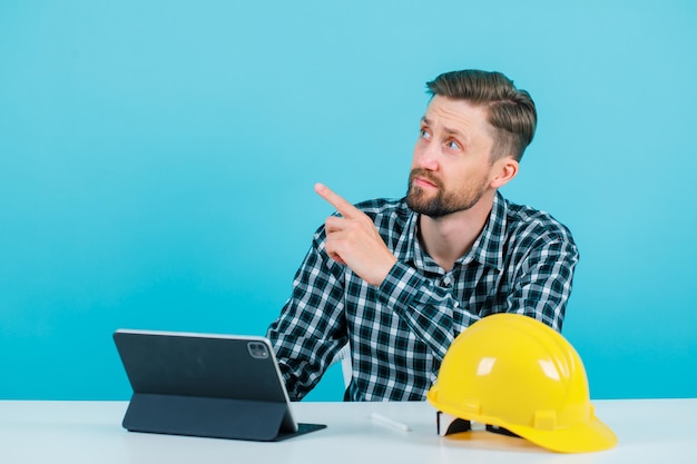 Foto grátis olhando para cima o homem de engenheiro está apontando para cima com o dedo indicador sentado na frente do tablet no fundo azul