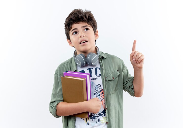 Foto grátis olhando para cima, o menino impressionado usando fones de ouvido, segurando o livro e aponta para cima, isolado no fundo branco com espaço de cópia