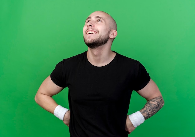 Foto grátis olhando para cima sorridente jovem desportivo usando pulseira e colocando as mãos no quadril sobre fundo verde