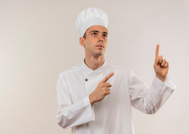 Olhando para o jovem cozinheiro usando uniforme de chef, aponta os dedos em direções diferentes na parede branca isolada com espaço de cópia