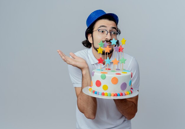 Olhando para o lado confuso de um homem bonito de óculos e chapéu azul, segurando um bolo e espalhando a mão isolada no fundo branco
