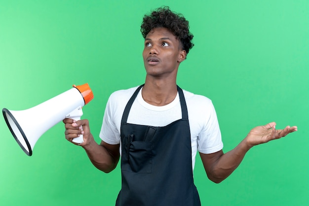 Olhando para o lado confuso, jovem barbeiro afro-americano vestindo uniforme, segurando o alto-falante e espalhando a mão isolada sobre fundo verde