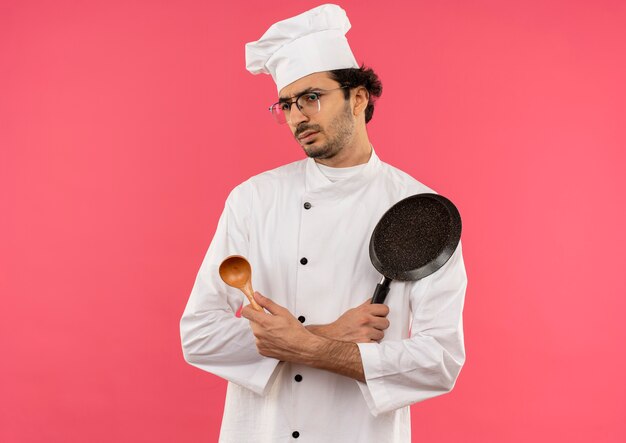 Olhando para o lado confuso, jovem cozinheiro vestindo uniforme de chef e óculos segurando e cruzando a colher com a frigideira