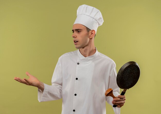 Olhando para o lado confuso, jovem cozinheiro vestindo uniforme de chef segurando a frigideira na parede verde isolada