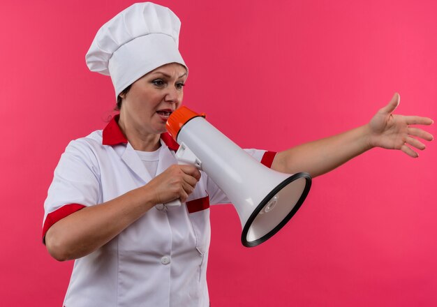 Olhando para o lado, cozinheira de meia-idade em uniforme de chef falando no alto-falante na parede rosa isolada