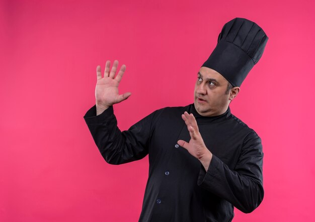 Olhando para o lado, cozinheiro masculino de meia-idade com uniforme de chef, mostrando o tamanho na parede rosa isolada com espaço de cópia