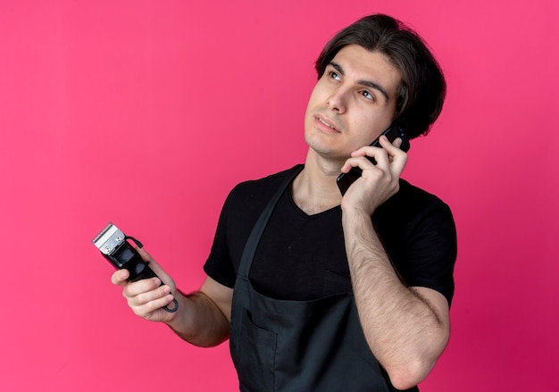 olhando para o lado de um jovem bonito barbeiro de uniforme segurando uma tesoura de cabelo e falando ao telefone