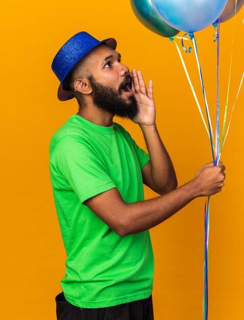 Olhando para o lado, jovem afro-americano com chapéu de festa segurando balões chamando alguém isolado na parede laranja