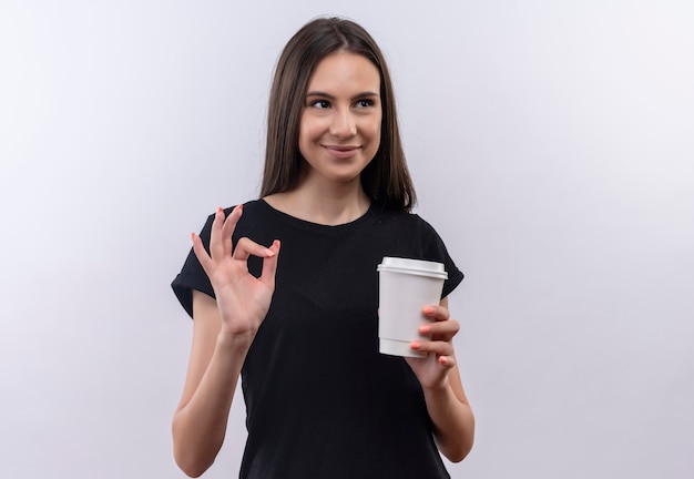 Olhando para o lado, jovem caucasiana, vestindo uma camiseta preta segurando uma xícara de café, mostrando um gesto de aprovação no fundo branco isolado