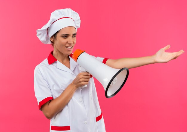 Olhando para o lado, jovem cozinheira vestindo uniforme de chef, fala no alto-falante apontando lado a lado na parede rosa isolada com espaço de cópia