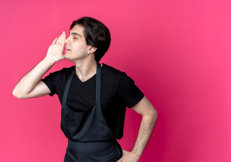 Foto grátis olhando para o lado, jovem e bonito barbeiro de uniforme sussurra e coloca a mão no quadril isolado na parede rosa com espaço de cópia