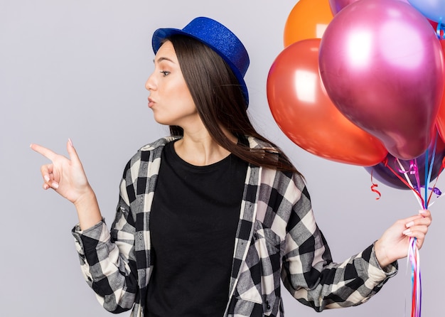 Olhando para o lado, jovem e linda garota usando chapéu de festa segurando balões apontando ao lado