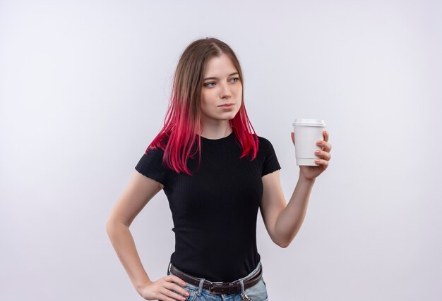 Olhando para o lado, jovem e linda garota vestindo uma camiseta preta segurando a xícara de café, colocando a mão no quadril em um fundo branco isolado