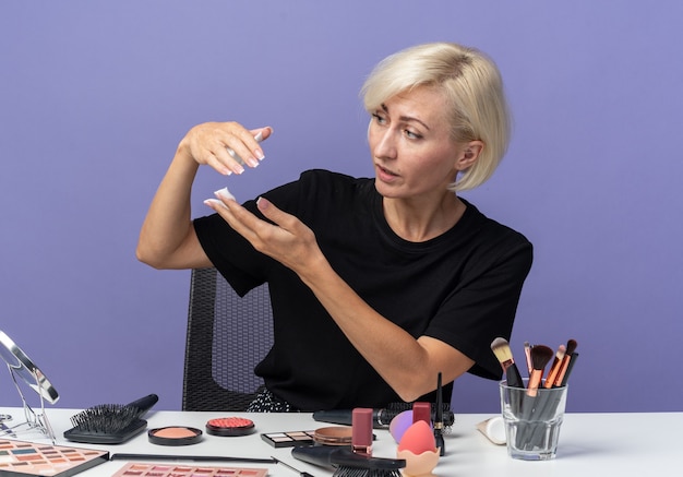 Olhando para o lado, jovem linda se senta à mesa com ferramentas de maquiagem, segurando creme para cabelo isolado na parede azul