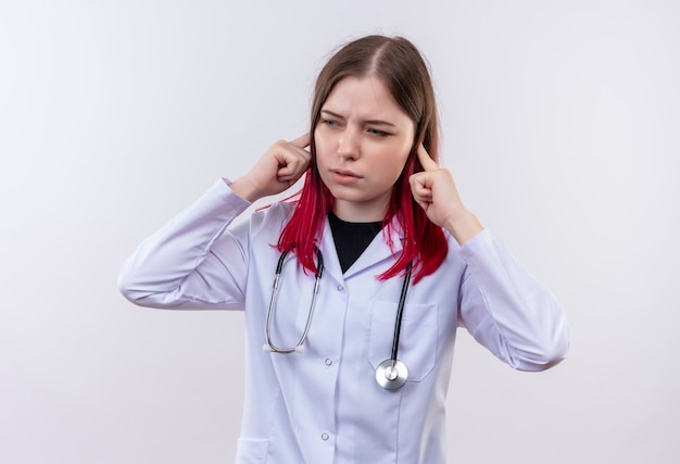 Olhando para o lado, jovem médica usando estetoscópio, manto médico, orelhas fechadas em fundo branco isolado
