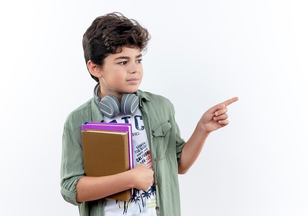 Foto grátis olhando para o lado, o menininho usando fones de ouvido, segurando um livro e apontando para o lado, isolado no fundo branco com espaço de cópia