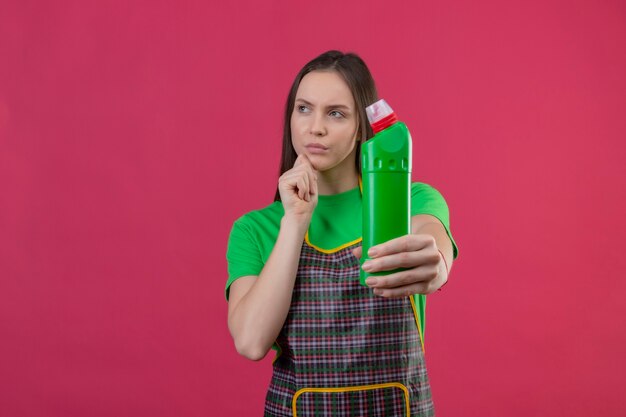 Olhando para o lado, pensando em limpar, jovem vestindo uniforme segurando um agente de limpeza com a mão no queixo na parede rosa isolada