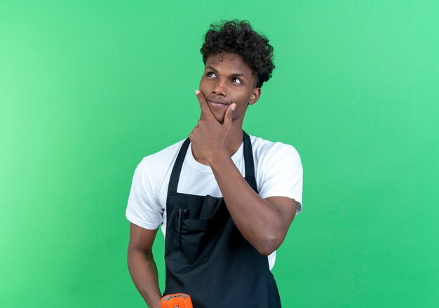 Olhando para o lado pensando jovem barbeiro afro-americano vestindo uniforme segurando o alto-falante e colocando a mão no queixo