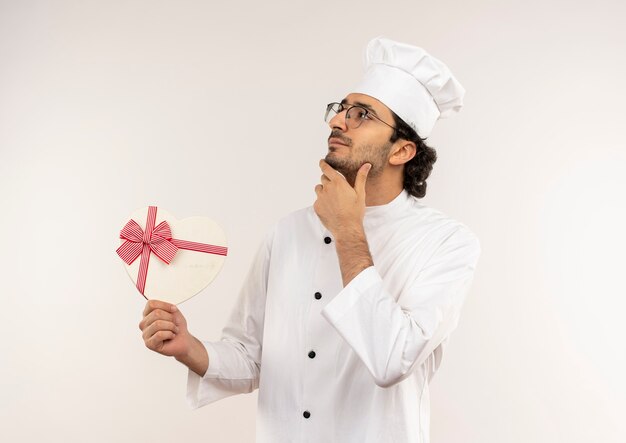 Olhando para o lado pensando jovem cozinheiro usando uniforme de chef e óculos segurando uma caixa de presente em forma de coração e colocando a mão no queixo