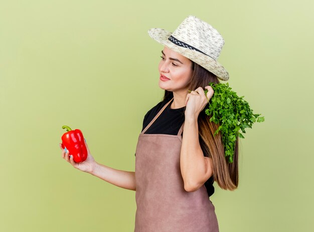 Olhando para o lado satisfeito, a linda garota jardineira de uniforme usando chapéu de jardinagem segurando pimenta com coentro isolado em fundo verde oliva