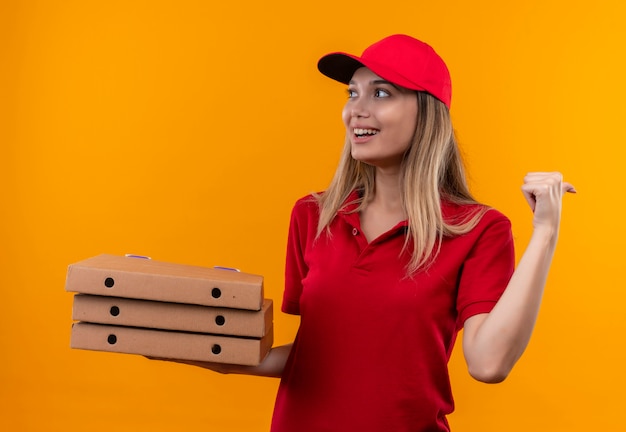 Olhando para o lado sorridente jovem entregador de uniforme vermelho e boné segurando uma caixa de pizza e pontos para o lado isolado em fundo laranja