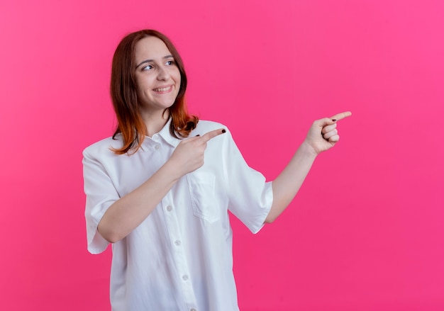 Olhando para o lado sorrindo, a jovem ruiva aponta para o lado isolado em um fundo rosa com espaço de cópia