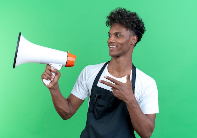 Olhando para o lado sorrindo, jovem barbeiro afro-americano, vestindo uniforme, segurando o alto-falante e mostrando gesto de pistola isolado no fundo verde