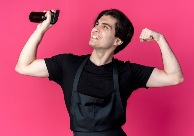 Olhando para o lado sorrindo jovem bonito barbeiro de uniforme segurando o frasco de spray e fazendo um gesto forte