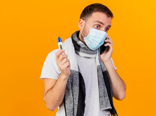 Olhando para o lado surpreso, jovem doente usando chapéu de inverno e máscara médica fala ao telefone e segurando um termômetro isolado em fundo amarelo