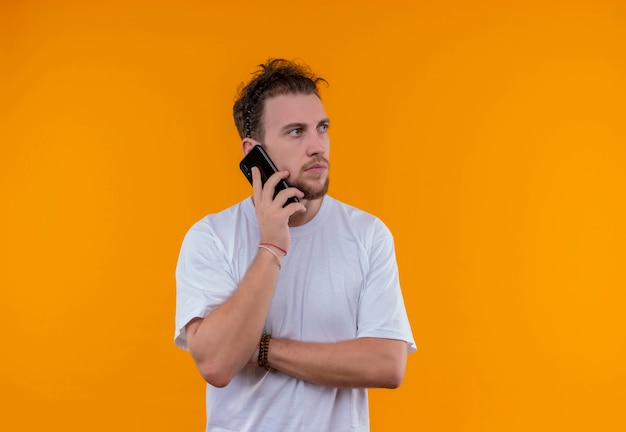 Olhando para o lado, um rapaz de camiseta branca fala ao telefone e cruza as mãos em um fundo laranja isolado