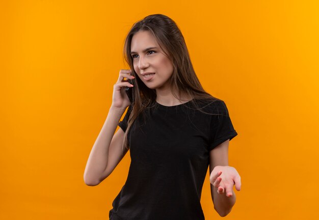 Olhando para o lado, uma jovem caucasiana de camiseta preta fala ao telefone em um fundo laranja isolado