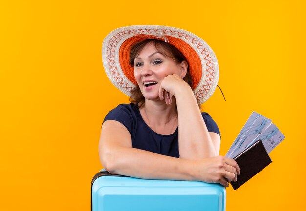 Olhando para o lado, uma mulher viajante de meia-idade com um chapéu segurando ingressos e uma carteira, colocando a mão na mala na parede amarela