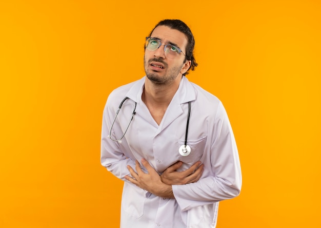 Foto grátis olhando para um jovem médico doente com óculos de médico, usando roupão médico com estetoscópio na barriga