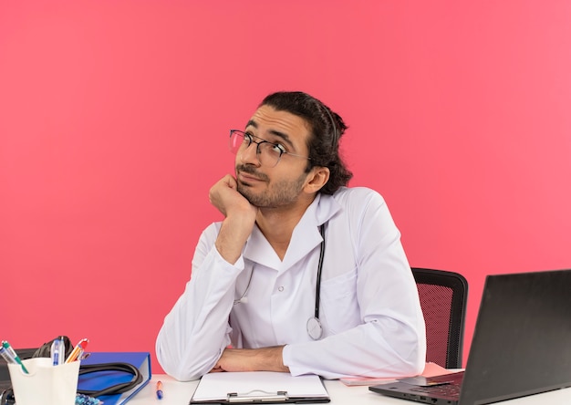 Olhando para um jovem médico satisfeito com óculos de médico, vestindo bata médica e estetoscópio sentado na mesa