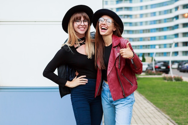 Olhar de moda outono. Duas garotas graciosas atraentes em giros óculos redondos e chapéus pretos, posando no centro de negócios.