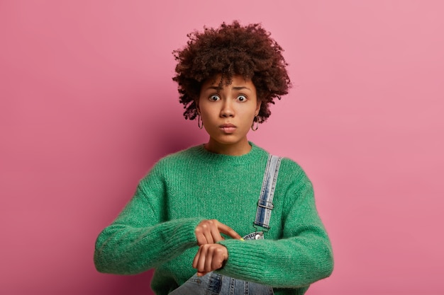 Foto grátis olhe a hora. modelo feminino encaracolado de pele escura preocupado aponta para o pulso, intrigado com o prazo, estar atrasado, parece nervoso, vestido de suéter verde, tem expressão envergonhada, posa dentro de casa