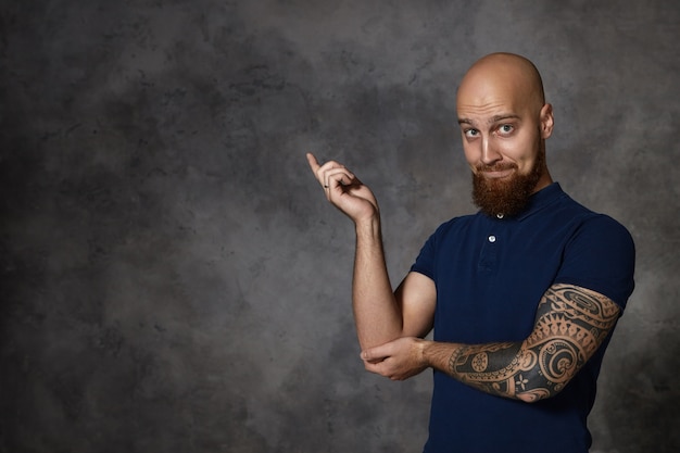 Olhe para isso! homem barbudo careca engraçado isolado com tatuagem levantando o dedo indicador e apontando para o canto esquerdo, expressando entusiasmo ou curiosidade, levantando as sobrancelhas. linguagem corporal