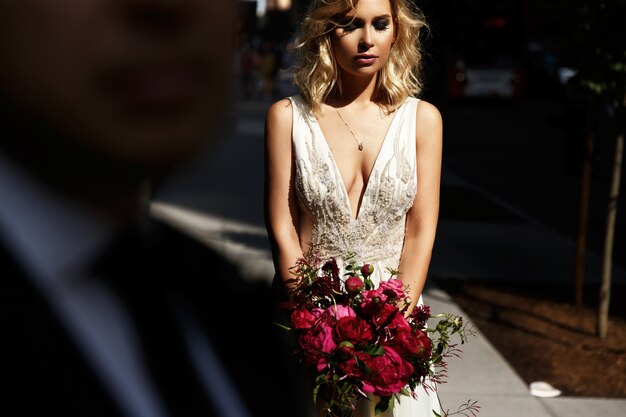 Olhe sobre o ombro do noivo na noiva de pé com o bouquet de casamento vermelho com luz do sol brilhante