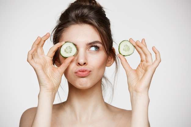 Olho escondido de sorriso da menina despida bonita nova atrás da fatia do pepino sobre o fundo branco. Conceito de spa e cosmetologia de beleza.