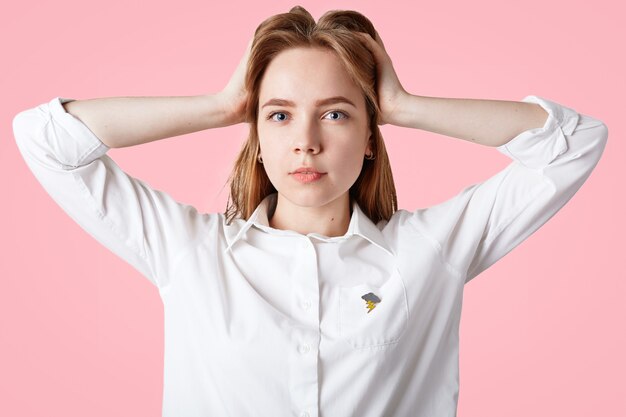 olhos azuis jovem fêmea com cabelos longos, mantém as mãos na cabeça e parece sério, vestido com camisa branca elegante, posa em rosa. Pessoas, expressões faciais, beleza