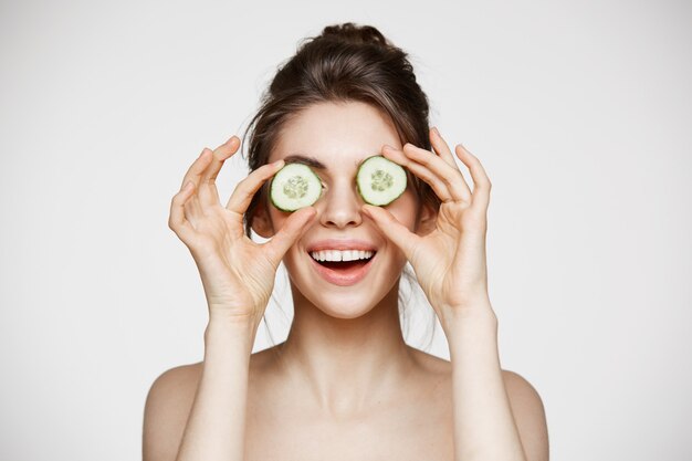 Olhos escondendo de sorriso da menina bonita nova do nude atrás das fatias do pepino sobre o fundo branco. Cuidados com a pele e cosmetologia.