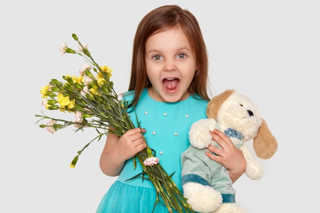 Onjoyed criança pequena de olhos azuis mantém seu brinquedo favorito e flores, feliz em receber presente no aniversário, abre a boca amplamente, vestida com vestido de festa, isolado no branco