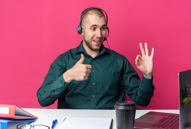 Operador de call center jovem sorridente usando fone de ouvido, sentado na mesa com ferramentas de escritório, olhando para o laptop, mostrando o polegar para cima e um gesto de ok