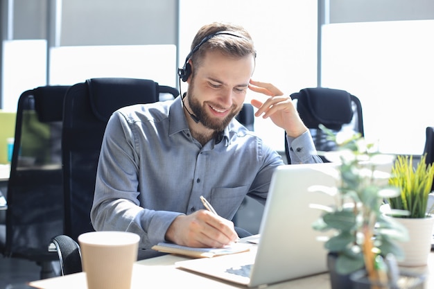 Operador de call center masculino sorridente com fones de ouvido sentado em um escritório moderno, consultando informações on-line em um laptop, procurando informações em um arquivo para ajudar o cliente.