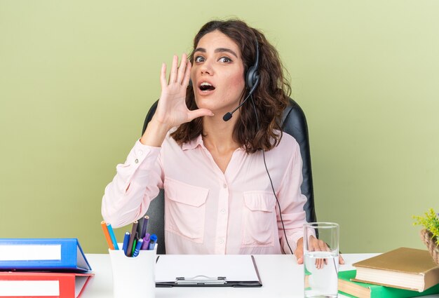 Operadora de call center do sexo feminino muito branca e surpresa com fones de ouvido, sentada na mesa, com ferramentas de escritório, mantendo a mão perto da boca