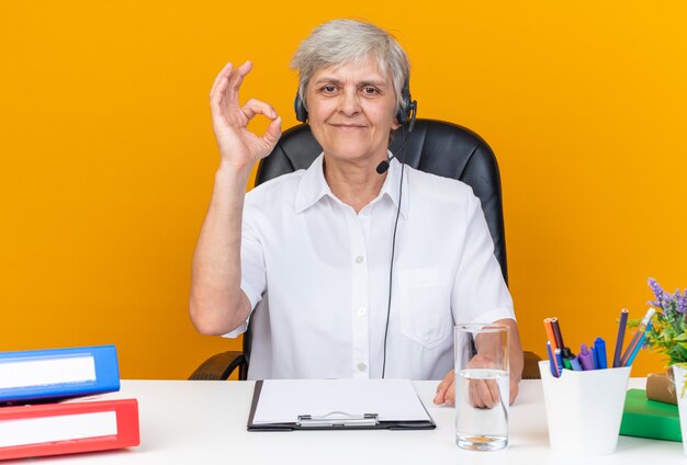 Operadora de call center feminina, caucasiana, satisfeita, com fones de ouvido, sentada na mesa com ferramentas de escritório, gesticulando sinal de ok