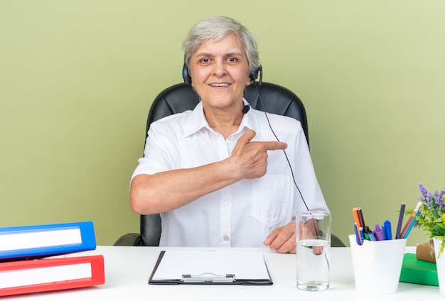 Operadora de call center feminina caucasiana sorridente em fones de ouvido, sentada na mesa com ferramentas de escritório apontando para o lado isolado na parede verde