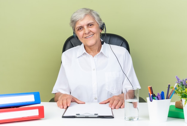 Operadora de call center feminina caucasiana sorridente em fones de ouvido, sentada na mesa com ferramentas de escritório, segurando uma prancheta isolada na parede verde
