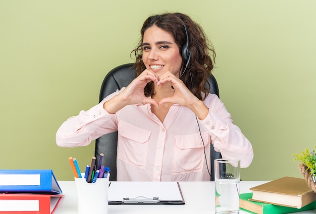 Operadora de call center feminina e bonita caucasiana em fones de ouvido, sentada na mesa com ferramentas de escritório, gesticulando com o coração