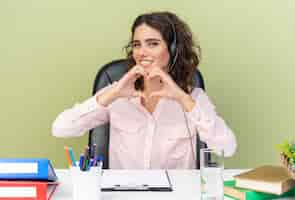 Foto grátis operadora de call center feminina e bonita caucasiana em fones de ouvido, sentada na mesa com ferramentas de escritório, gesticulando com o coração